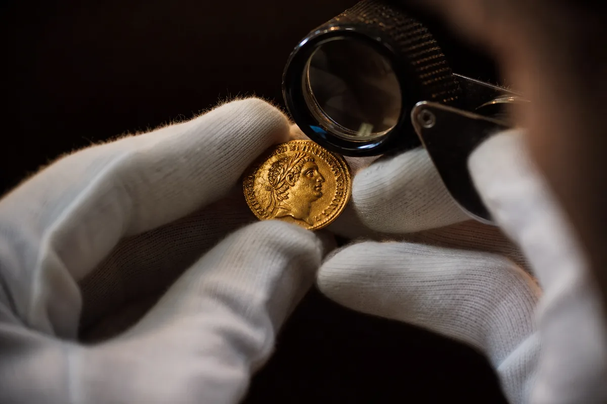 Mains gantées de coton blanc examinant une pièce d’or ancienne à la loupe de bijoutier sous une lumière chaude, fond sombre
